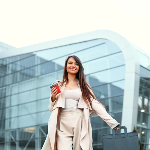Mulher de negócios sorridente caminhando em frente a um edifício moderno de vidro, segurando um copo de café e puxando uma mala de mão executiva.