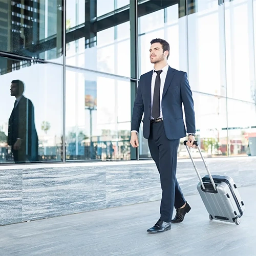 Homem de negócios vestindo terno azul-marinho caminha em frente a uma fachada de vidro moderna, puxando uma mala de rodinhas prateada.