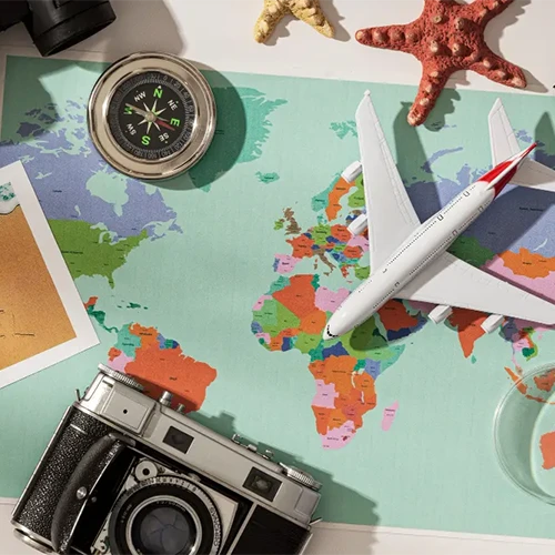 Visão aérea de itens de viagem sobre um mapa-múndi colorido, incluindo uma câmera fotográfica analógica prateada, uma bússola de metal, estrelas-do-mar e uma miniatura de avião branco.
